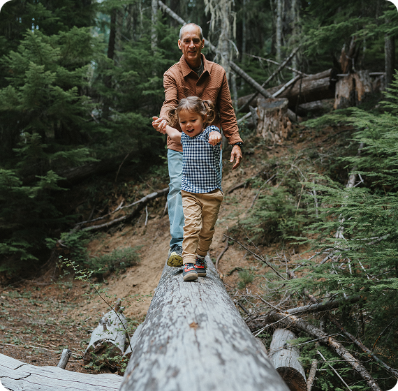 Image of grandparent helping a child balance while walking on a fallen log in the woods.