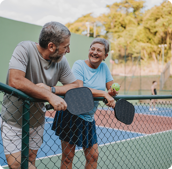 Image of two people smiling and talking while holding pickleball paddles by a court.