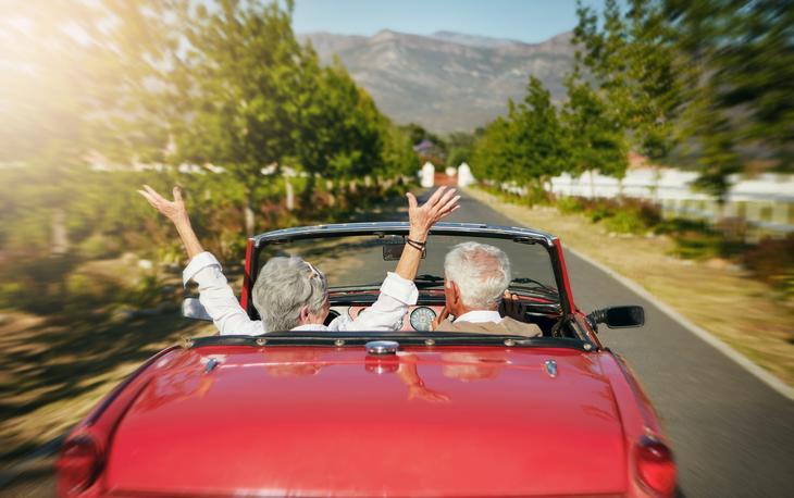 A retired couple in a red convertible happily driving down a road on a sunny day.